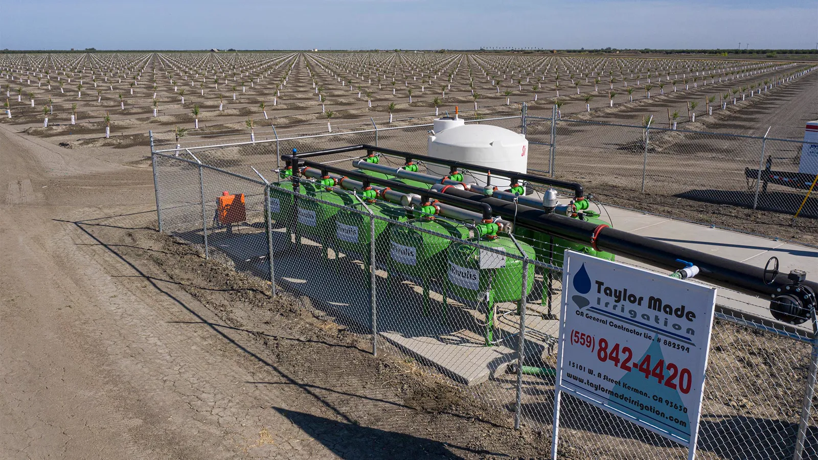 Green sand media filter tanks inside a chain-link fence beside young almond rows, with the Taylor Made Irrigation job sign wired to the fence.