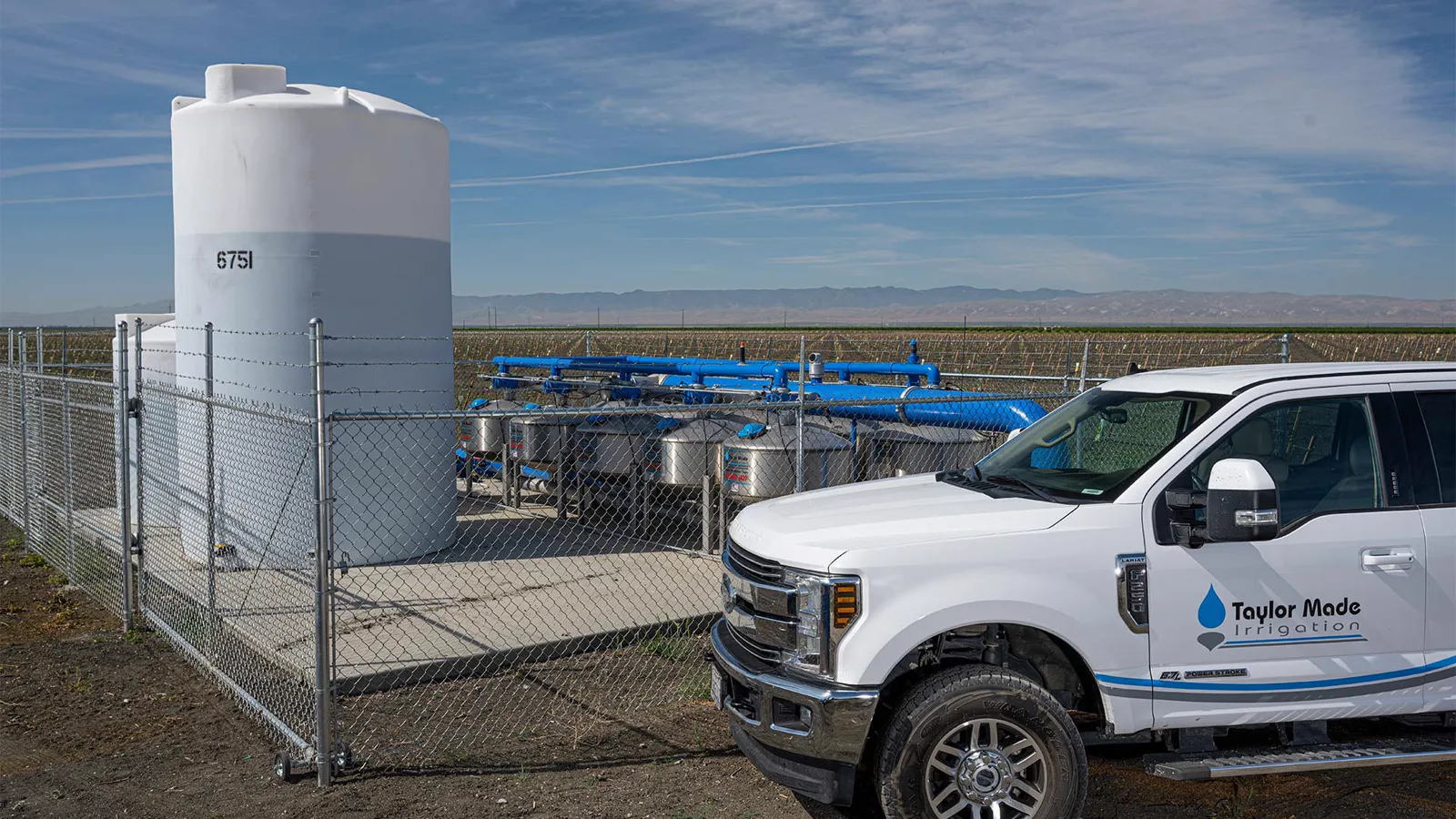 A white Taylor Made Irrigation pickup, lettered with the blue drop, parked beside a fenced filter station at Holland Farms: stainless sand media tanks under blue manifold pipe, a tall white tank, young trees and hills on the horizon.