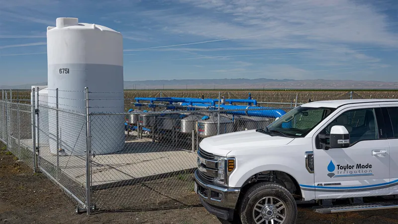 A white Taylor Made Irrigation pickup beside the fenced filter station at Holland Farms, blue manifold pipe over stainless tanks.
