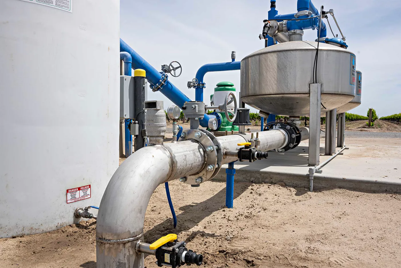At Pacheco Partnership, a welded stainless pipe elbow with bolted flanges and yellow-handled valves runs from a white tank to stainless filter tanks with blue piping, vineyard rows behind.